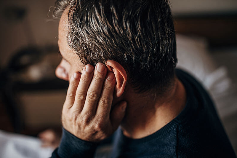 A man sits indoors, holding his hand to his ear as if experiencing discomfort or pain.