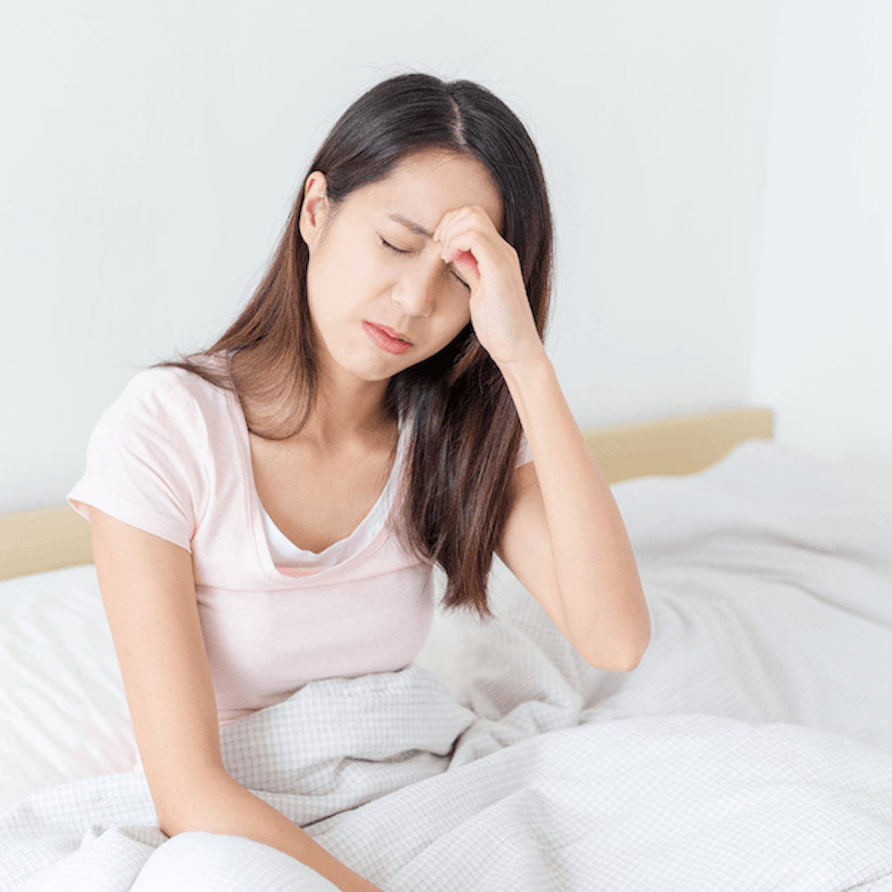 A woman sits on a bed with her eyes closed, holding her forehead with one hand, appearing to be in pain or discomfort.