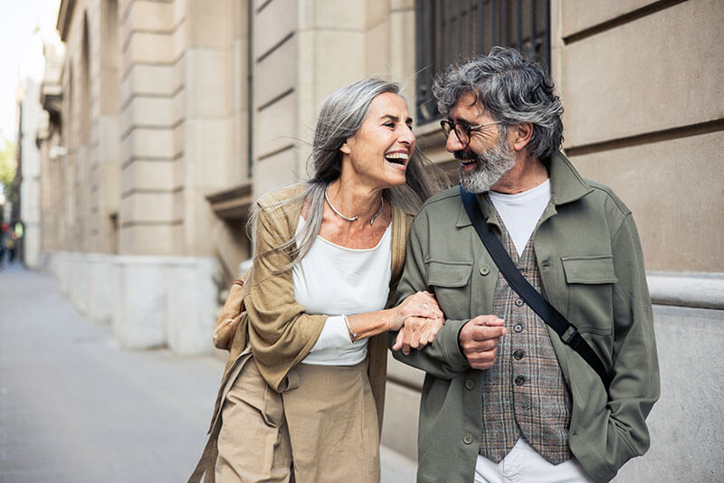 An older couple happily walks arm in arm down a city sidewalk, smiling and laughing together.