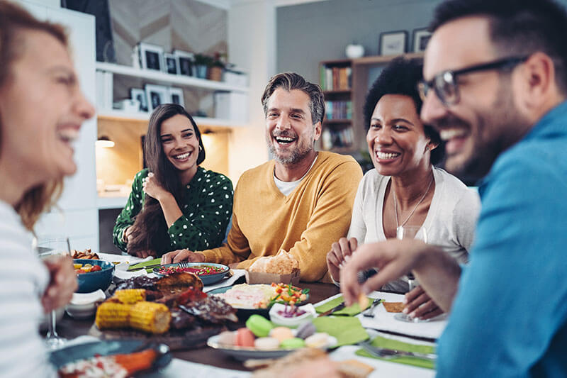 Five adults sit around a table, smiling and laughing while sharing a meal. The table is filled with various foods, including corn, grilled meat, and vegetables. The atmosphere appears warm and friendly.