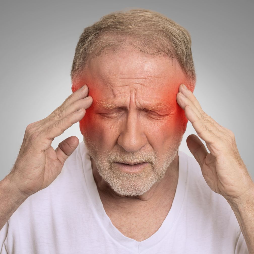 Older man with closed eyes and pained expression holding his temples; red coloring around his temples suggests a severe headache or migraine.