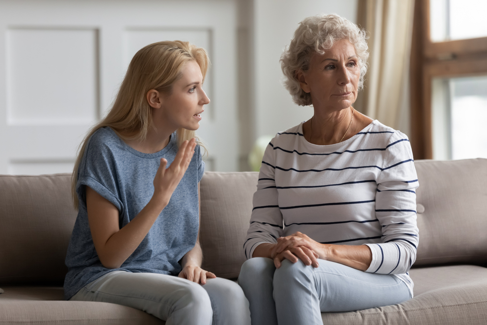 A young woman talks to an older woman on a couch. The older woman looks away with a serious expression, while the younger woman gestures with her hand, appearing concerned or upset.