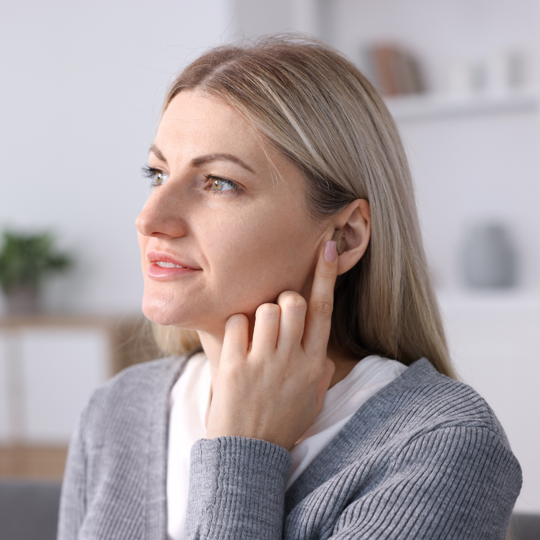 A woman with blonde hair touches her ear while looking to the side. She is wearing a gray sweater and sitting indoors in a softly lit room.