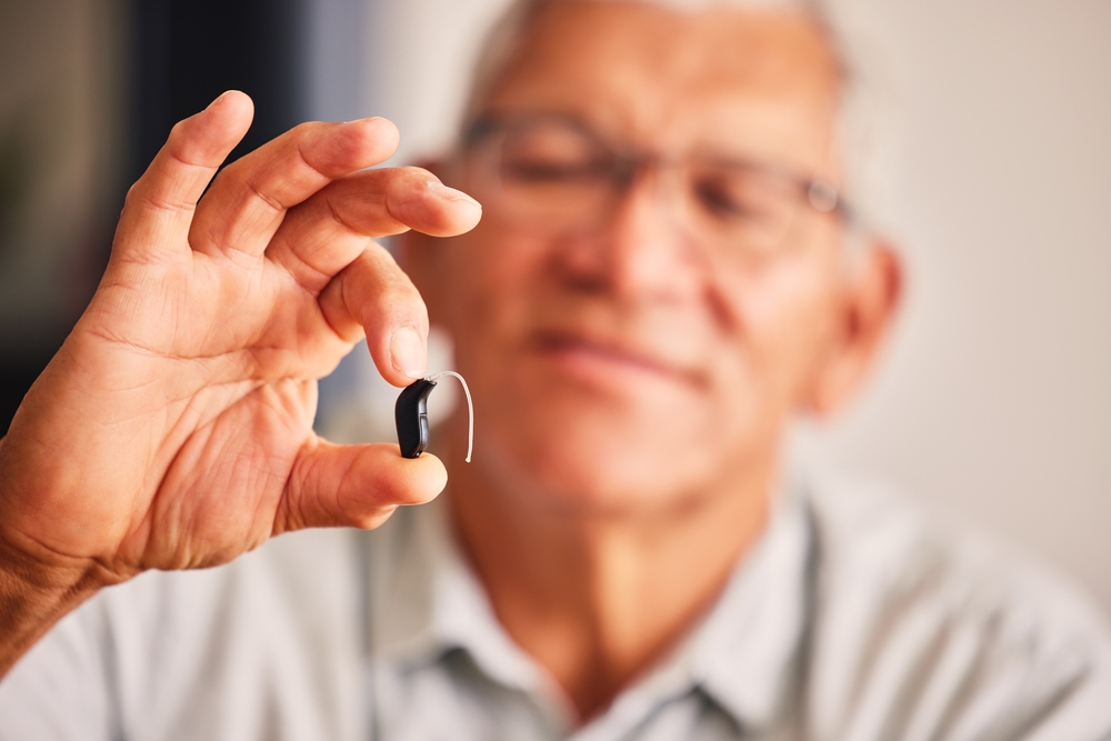 An older man holding a hearing aid between his fingers, smiling and looking at the device.